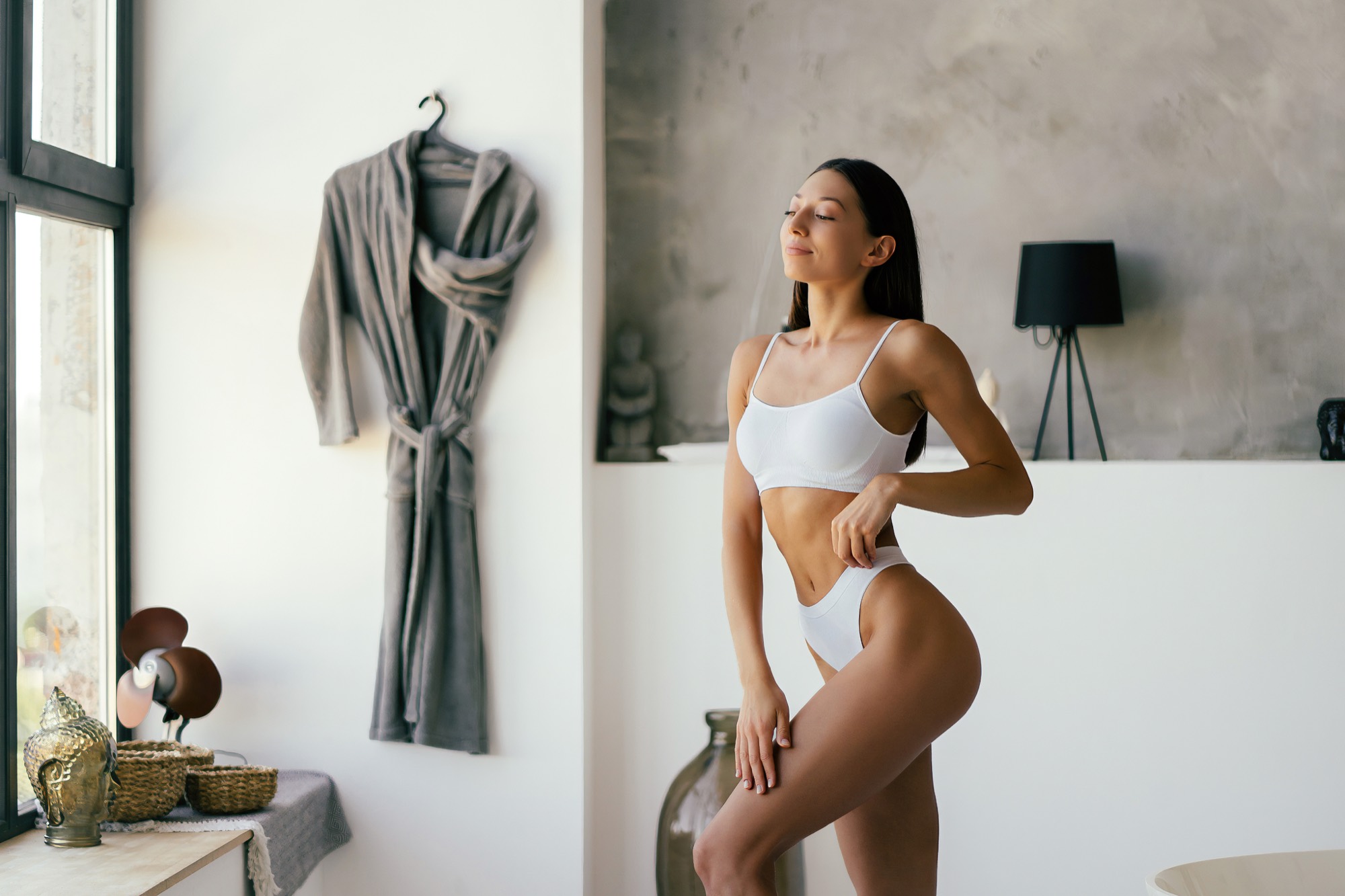 Woman in white underwear standing in a bright bedroom, one hand on her thigh